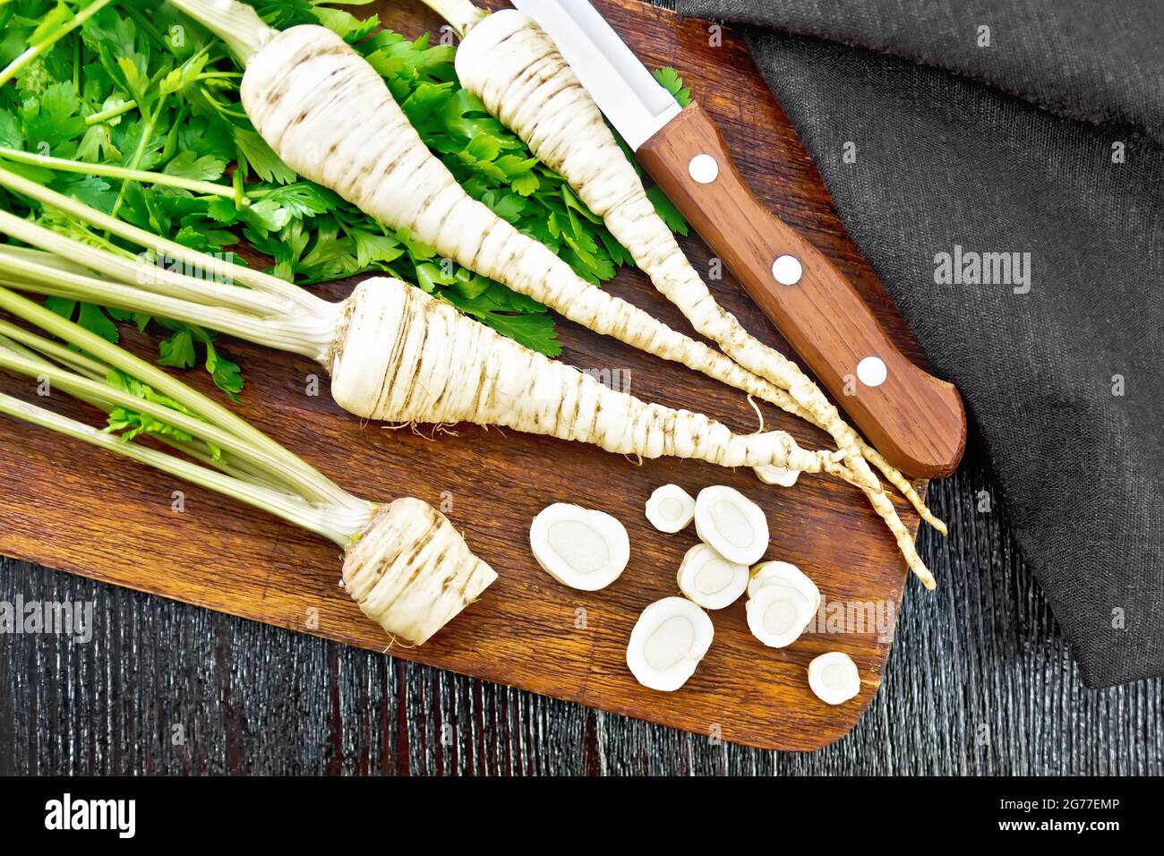 Parsley roots whole and chopped with green tops, knife, napkin on the background of a dark wooden board from above Stock Photo