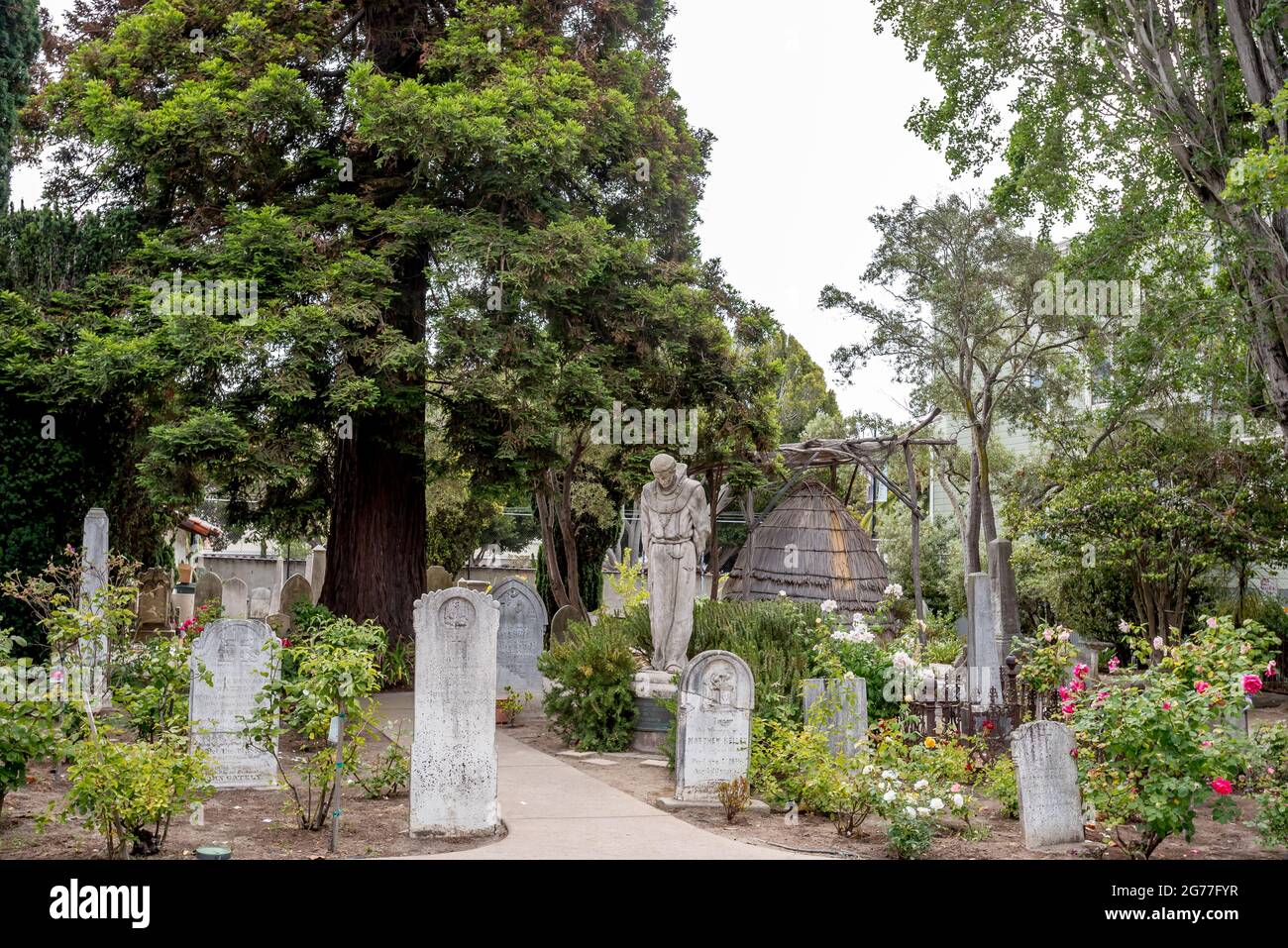 Cemetery at San Francisco Mission Dolores church. A statue of Father Junipero Serra stands in contrast with an Ohlone style indigenous family hut. Stock Photo