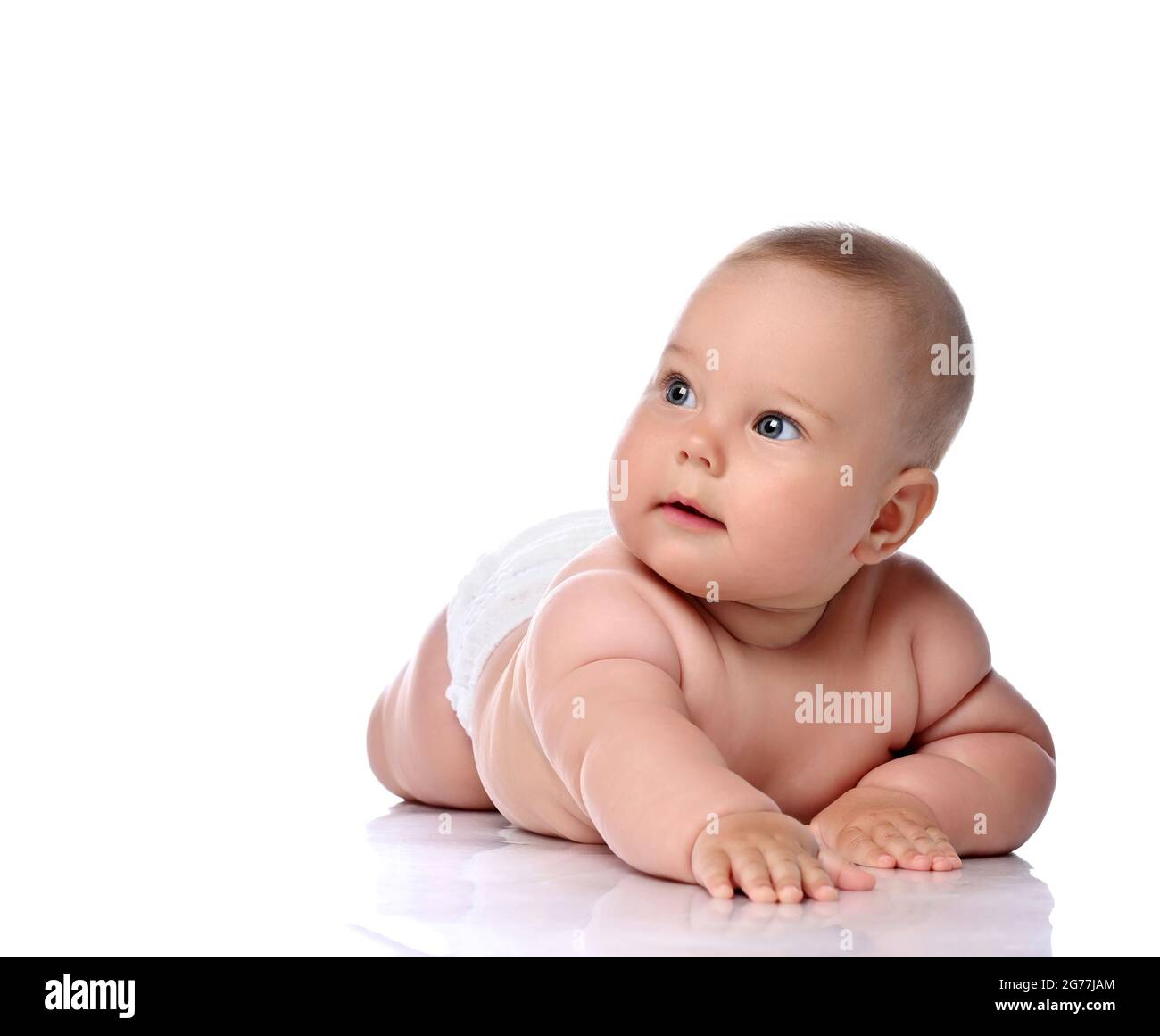 Surprised infant child baby girl kid in diaper is lying on her stomach slapping on floor and looking at upper corner  Stock Photo