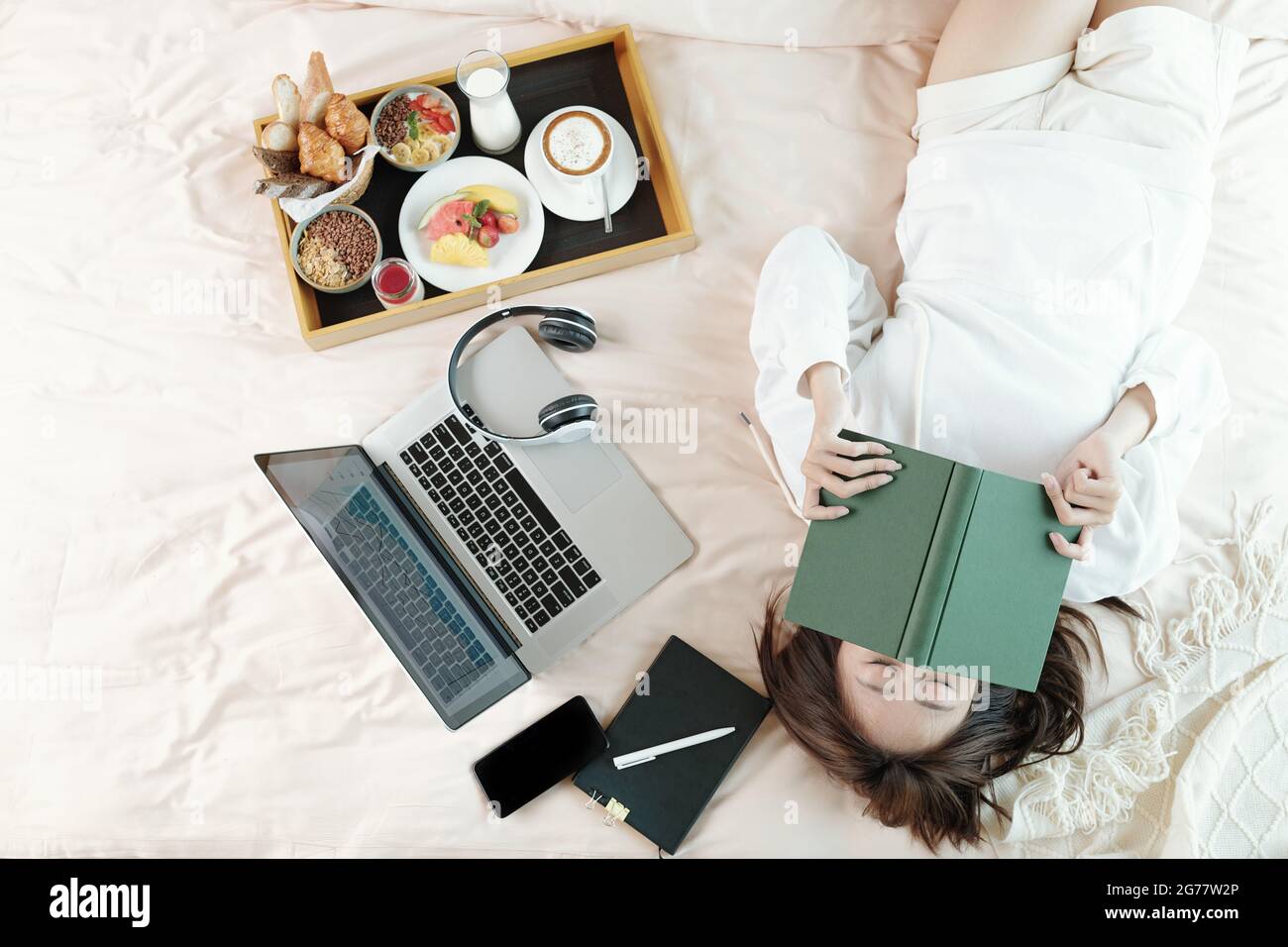 Lazy young woman lying on bed with opened laptop and tray with breakfast, sleeping after reading boring book Stock Photo