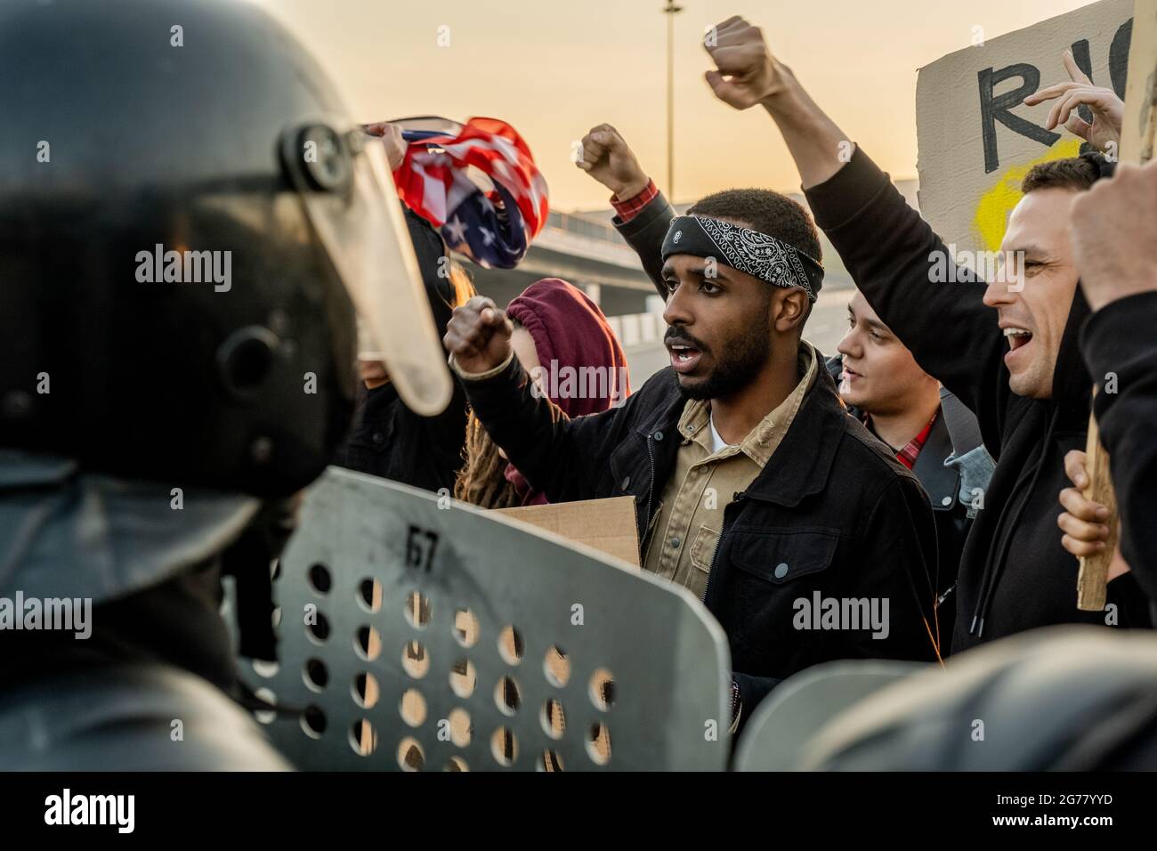 Group of disgruntled young multi-ethnic people raising arms and screaming protecting speech at riot while standing against police with shields Stock Photo