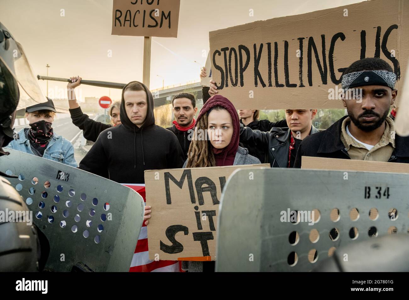 Group of serious young multi-ethnic activists standing with signs at street and protesting together, police guards stopping them with shields Stock Photo