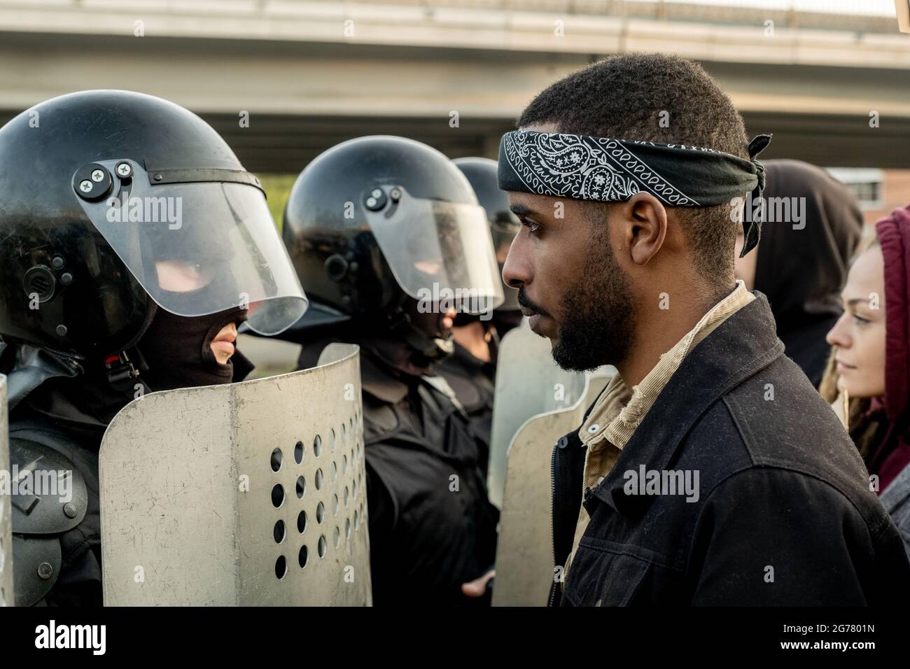 Disgruntled young Black bearded man in bandana standing in front of police officers with riot helmets and shields outdoors Stock Photo