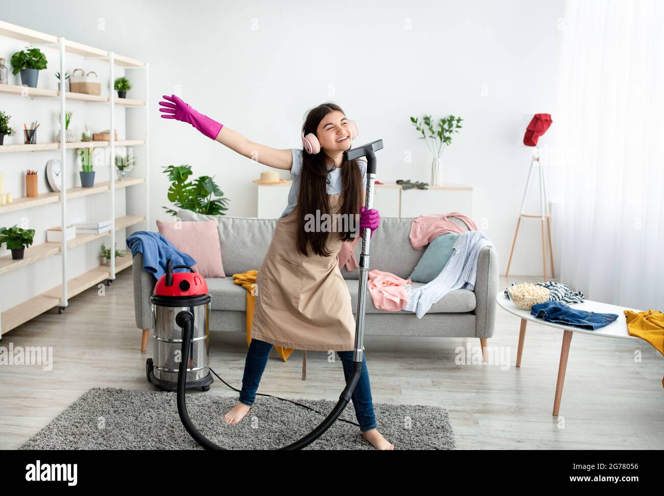 Happy Indian teen girl cleaning her home, wearing headphones, using vacuum cleaner as microphone and having fun Stock Photo