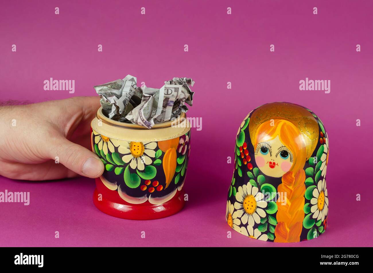 Hand holds Russian matryoshka full of money. Crumpled hundred dollar bills fall out of open wooden toy. Symbol of Russia and American money on pink ba Stock Photo