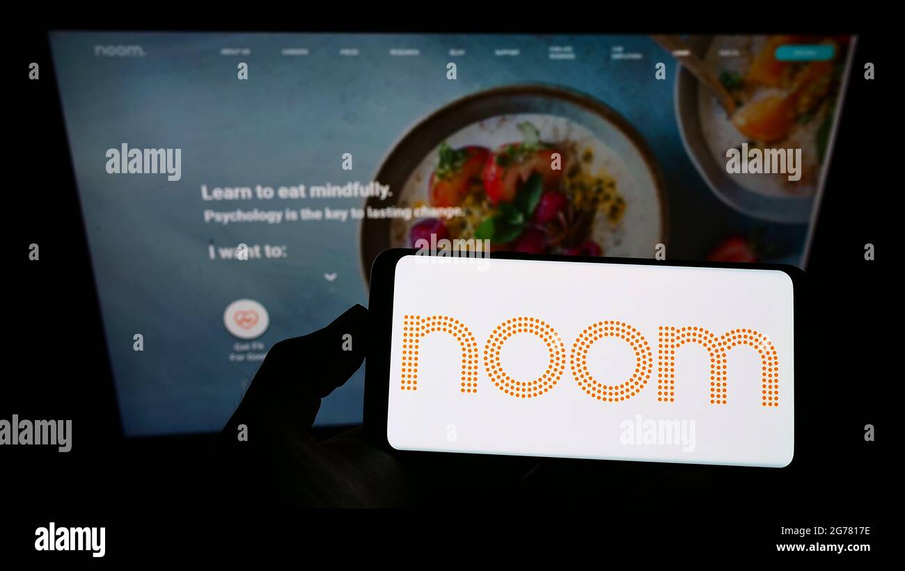 Person holding smartphone with logo of US weight-loss platform company Noom Inc. on screen in front of website. Focus on phone display. Stock Photo