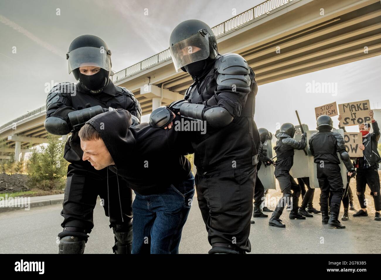 Riot police in body armors twisting man into uncomfortable position while confronting crowd in city Stock Photo