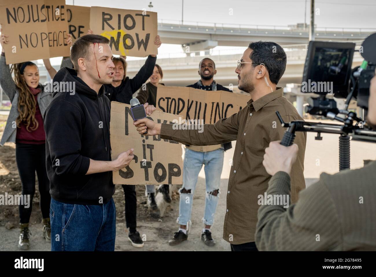 Young male rebel with broken head giving interview to Arabian journalist while other protestors waving banners in background Stock Photo