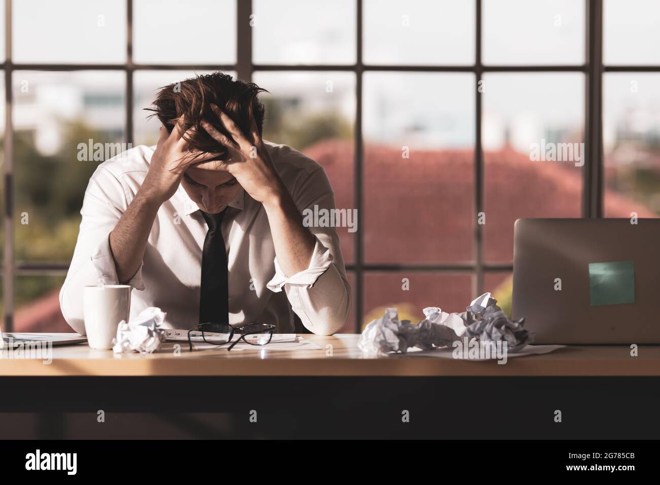 Young businessman sitting alone at desk and use hands holding his head feeling sad, serious and stress from work problems in office, there are surroun Stock Photo