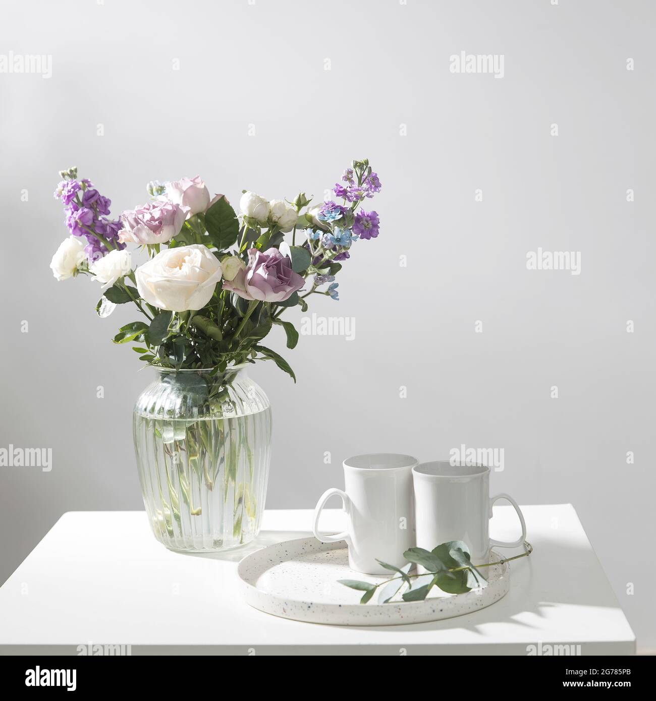 Bouquet of hackelia velutina, purple and white roses, small tea roses, matthiola incana and blue iris in glass vase is on the white coffee table with Stock Photo