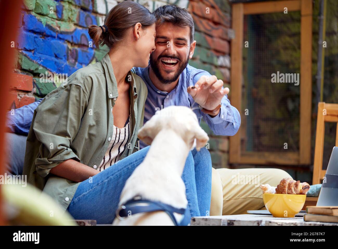 young caucasian couple having fun with their dog in outdoor cafe, feeding their pet, labrador retriever. Stock Photo