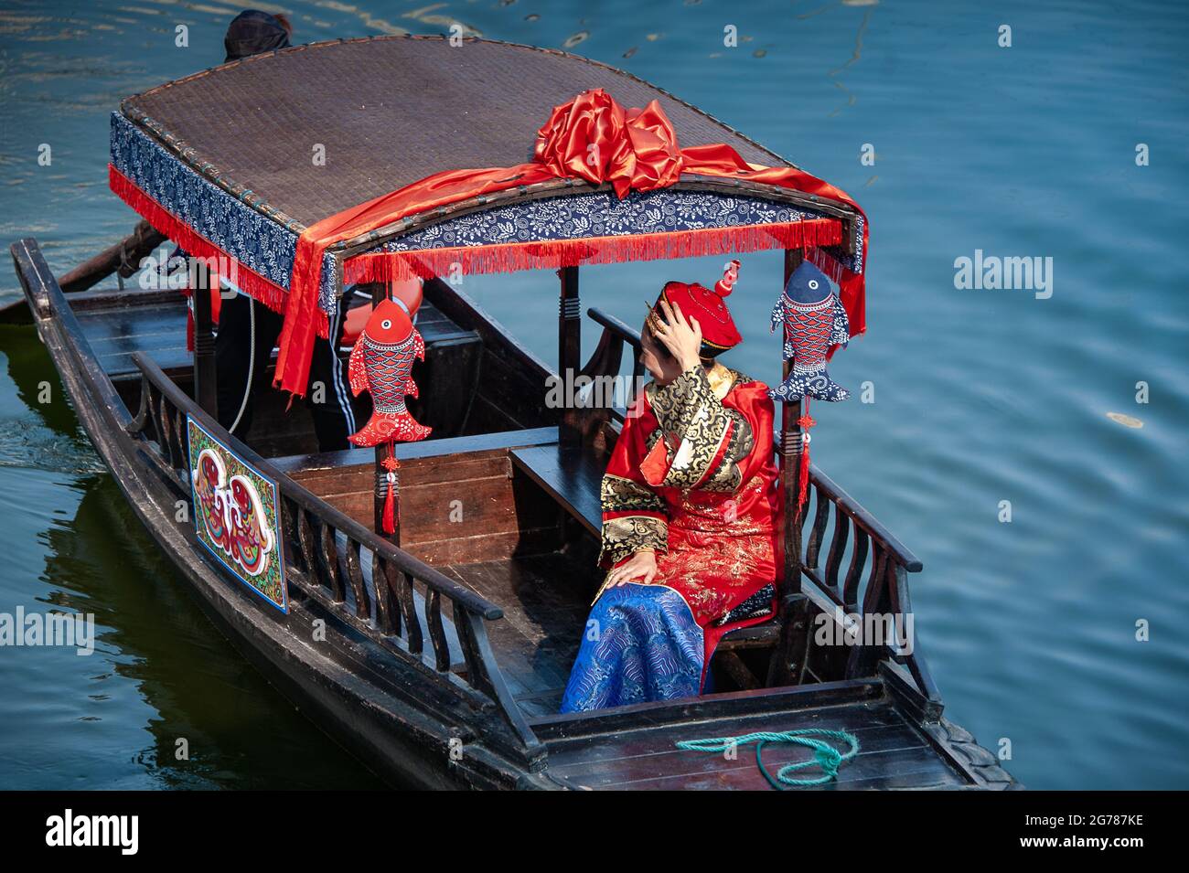 Wuxi, China - October 2019: Woman dressed in traditional Chinese wedding costume on a decorated river barge, against blue background. Stock Photo