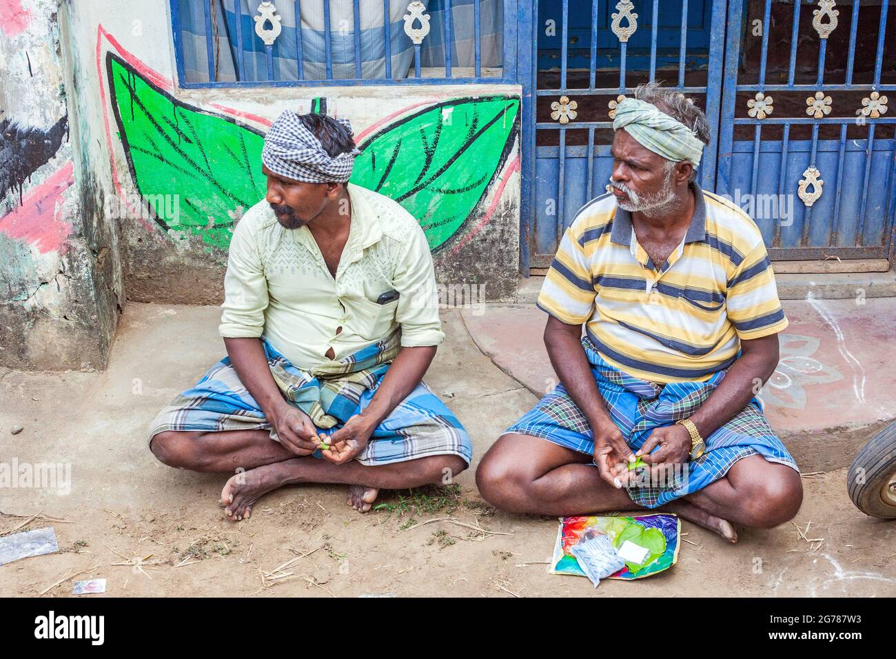 Two Indian male betel nut traders wearing lunghis seated on ground with betel leaf painted on wall behind them, Tamil Nadu, India Stock Photo