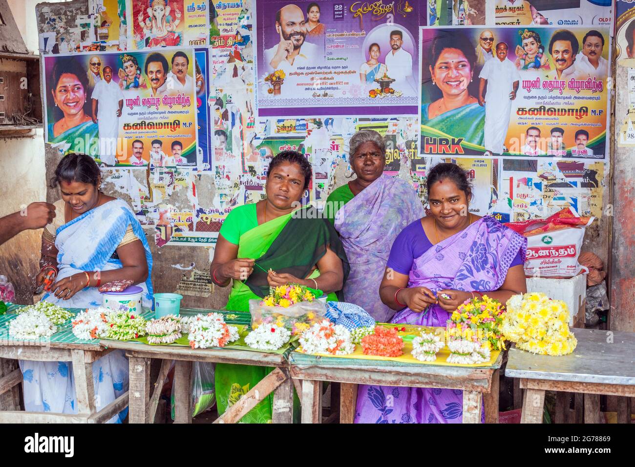 Indian females at market stall selling flower garlands as pooja temple offerings in front of colourful posters, Tamil Nadu, India Stock Photo