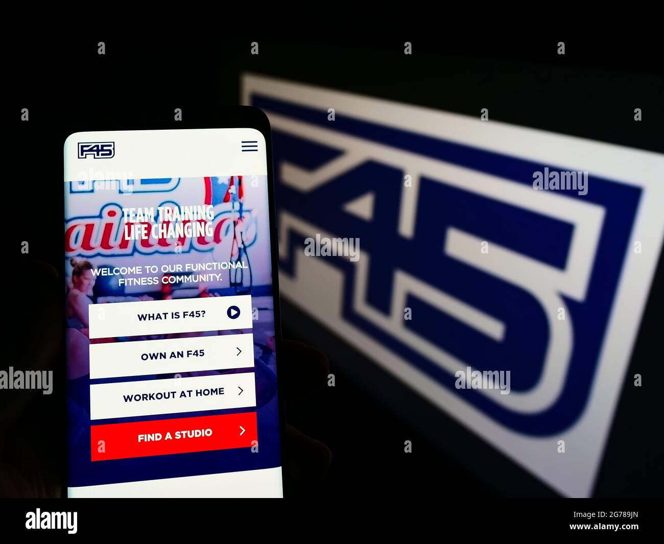 Person holding smartphone with website of fitness company F45 Training Holdings Inc. on screen in front of logo. Focus on center of phone display. Stock Photo