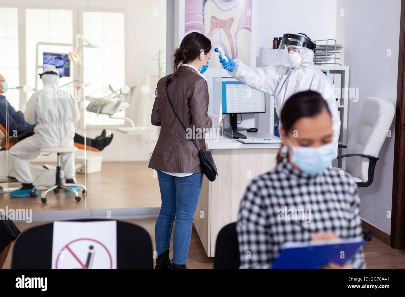 Stomatology receptionist using digital thermometer to measure patient temperature dressed in ppe suit as safety precaution during global outreabk with coronavirus. Dentist treating teeth. Stock Photo