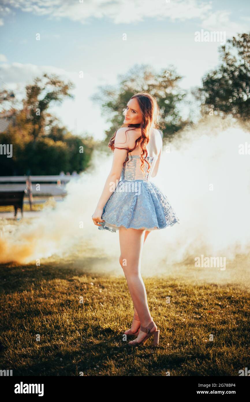 full-length portrait of a young woman in a short blue embroidered dress at a party in the park at sunset in summer with yellow smoke clouds on the Stock Photo