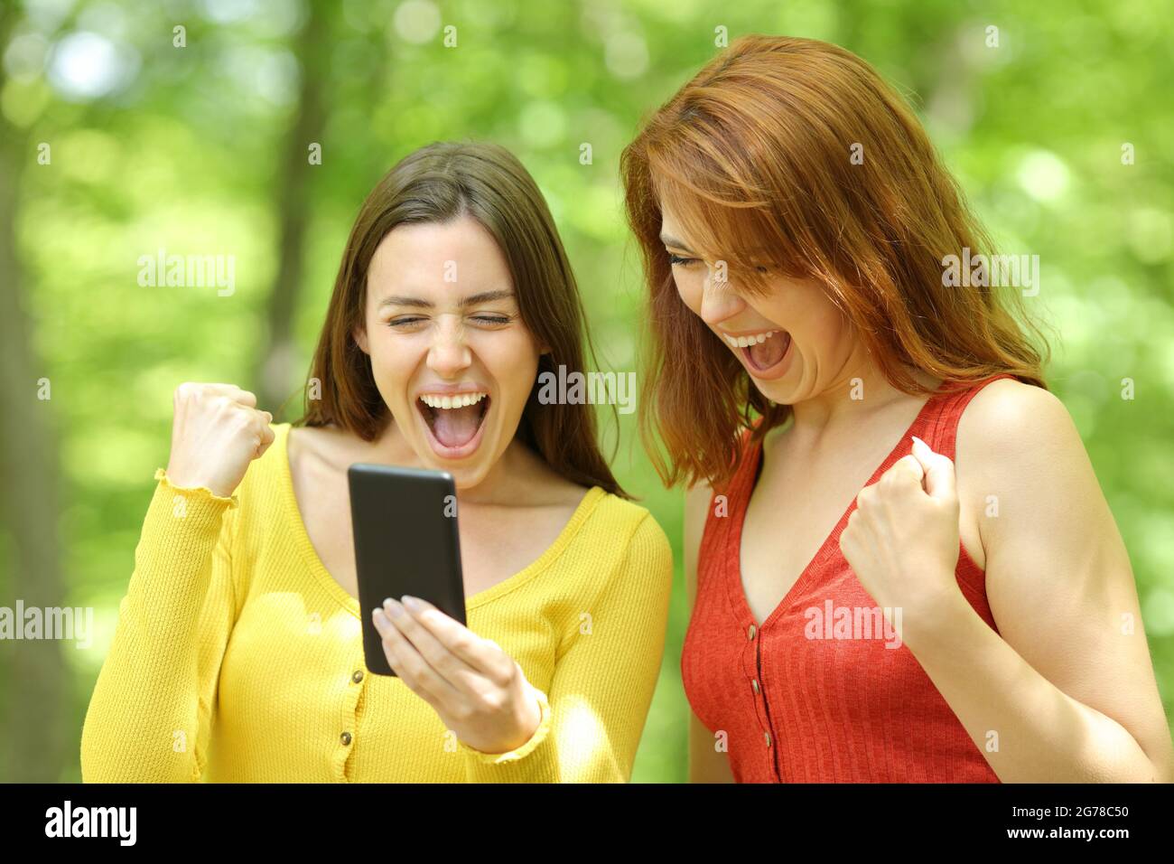 Two excited women checking smart phone content in a green park or forest Stock Photo