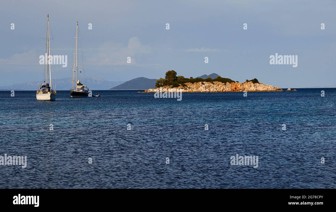 Ionian Islands, Ithaca, island of Odysseus, northeast coast, Marmakas Bay, two sailing boats anchor at the islet of Agios Nikolaos, the sea is dark blue, the sky is blue with clouds, behind the islet the island of Atokos rises Stock Photo