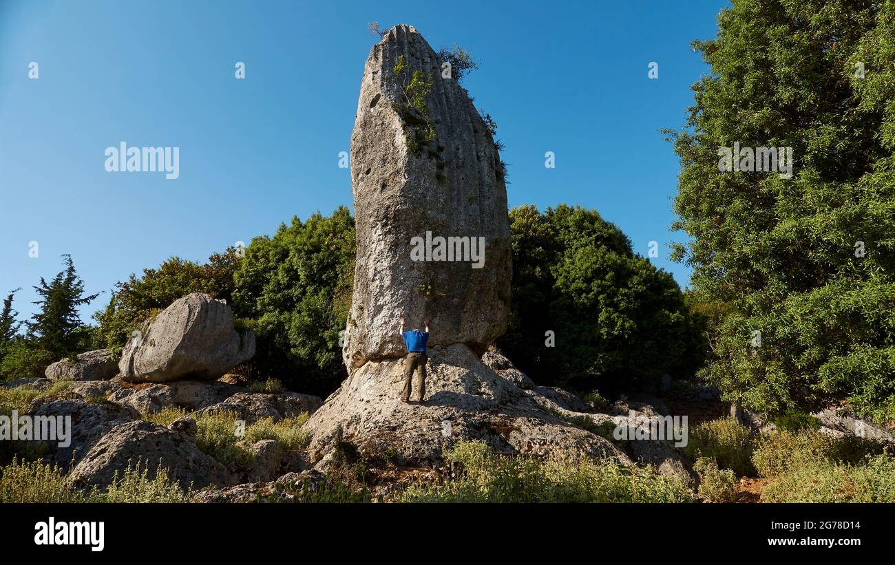Ionian Islands, Ithaka, Island of Odysseus, Anogi, Monolith of Anogi, Stone Monolith, 9m high, is called Araklis, man in a blue T-shirt braces against the monolith, trees next to and behind the monolith, blue sky above Stock Photo