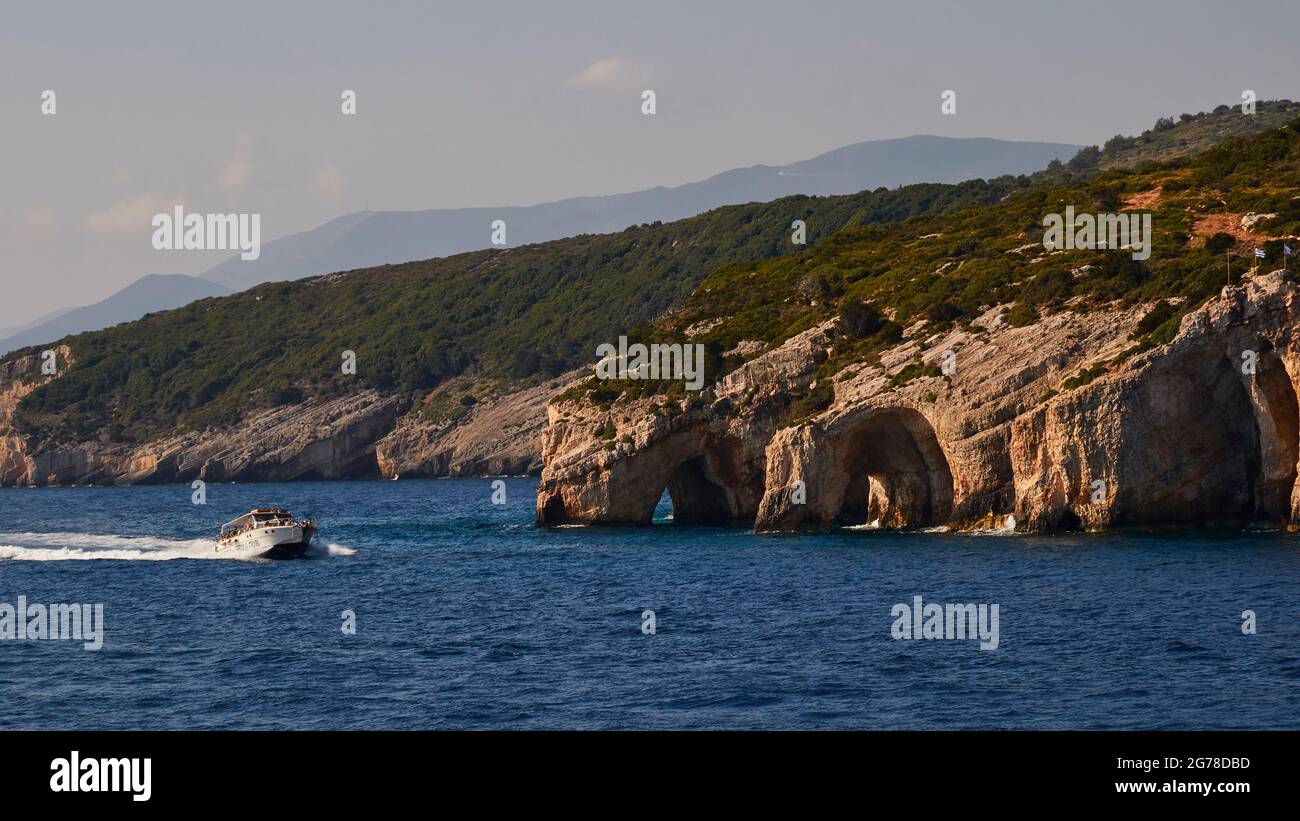 Ionian Islands, Zakynthos, northeast coast, Blue Caves, Blue Grottoes, morning light, view back to the north, excursion boat quickly drives into the picture, sky light blue to white, mountains in the haze Stock Photo
