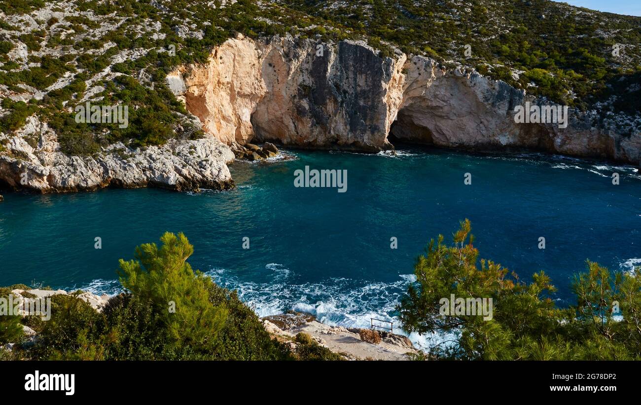 Ionian Islands, Zakynthos, west coast, Adriatic Sea, beach, harbor, Porto Limnionas, view from a little above over the bay, green bushes in the foreground, sea with white surf spray in the middle distance, green-blue water, overgrown rock face opposite to the top right, small blue streak of sky Stock Photo