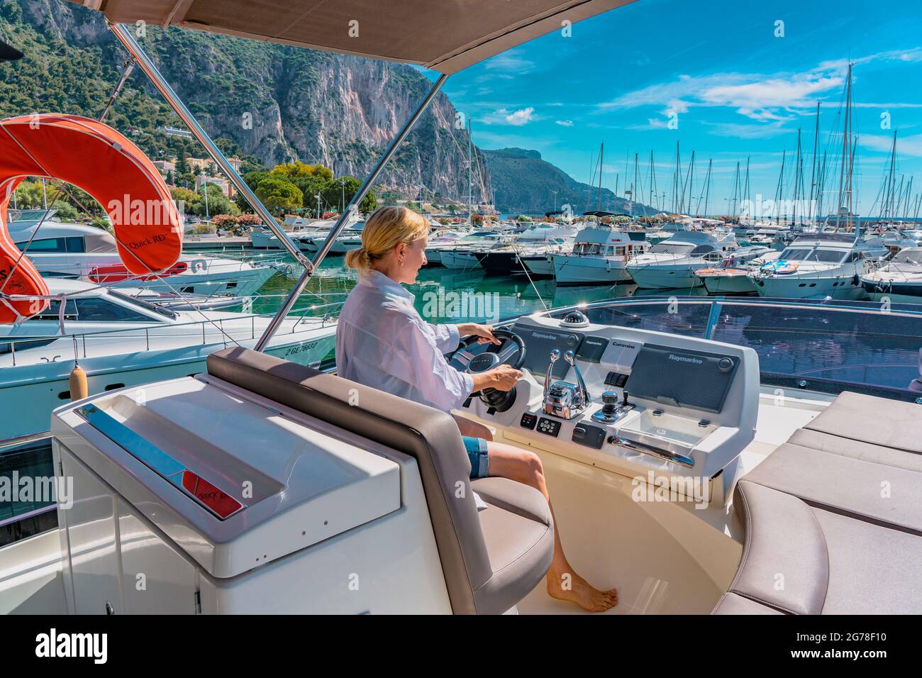 Beautiful blond woman driving yacht. Adult woman model in a white shirt sitting at the wheel of the yacht and begin to drive luxury boat from the port. Stock Photo