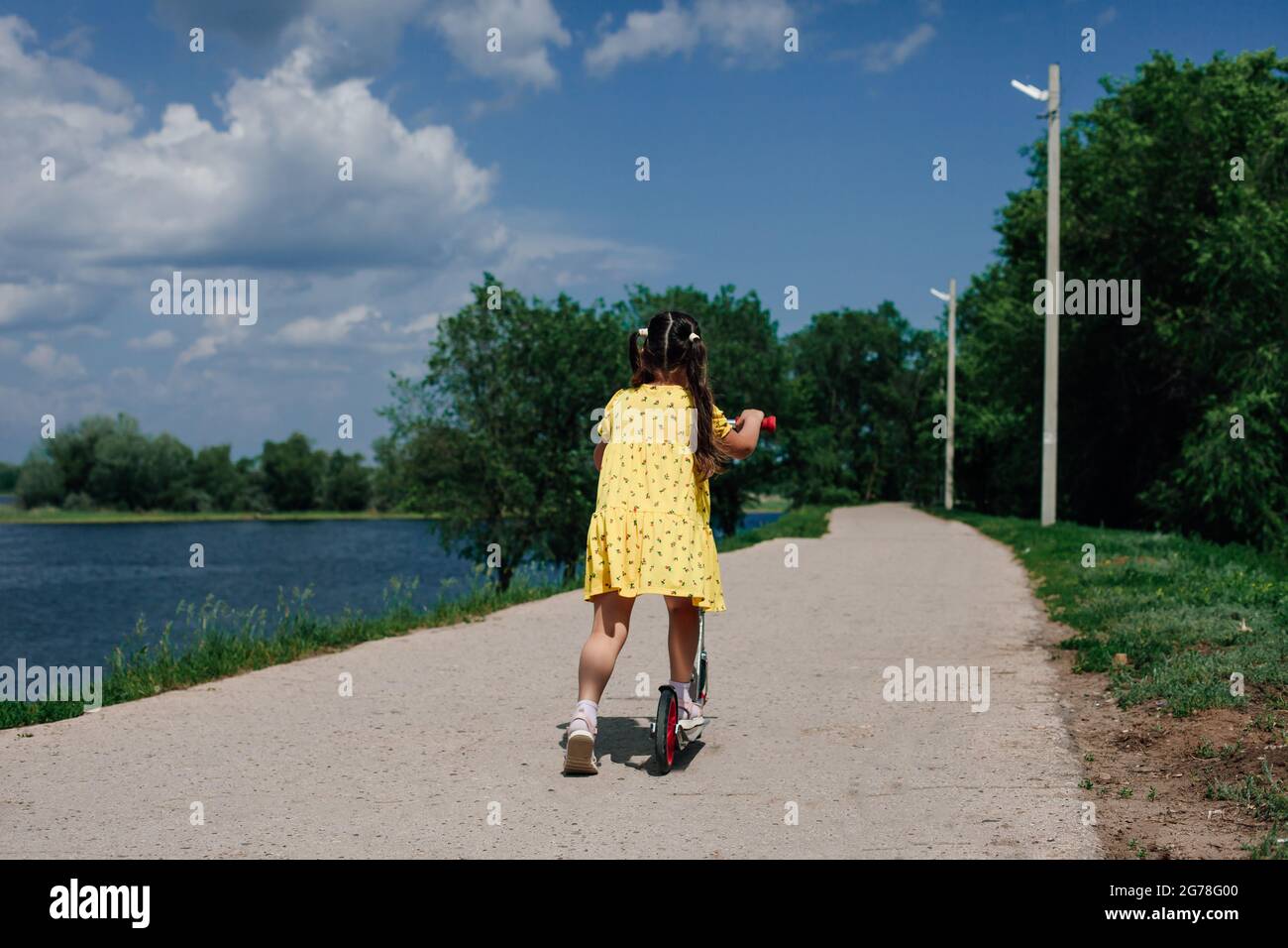 rear view of a girl in a yellow dress, a girl learns to ride a scooter in a natural park near the lake Stock Photo