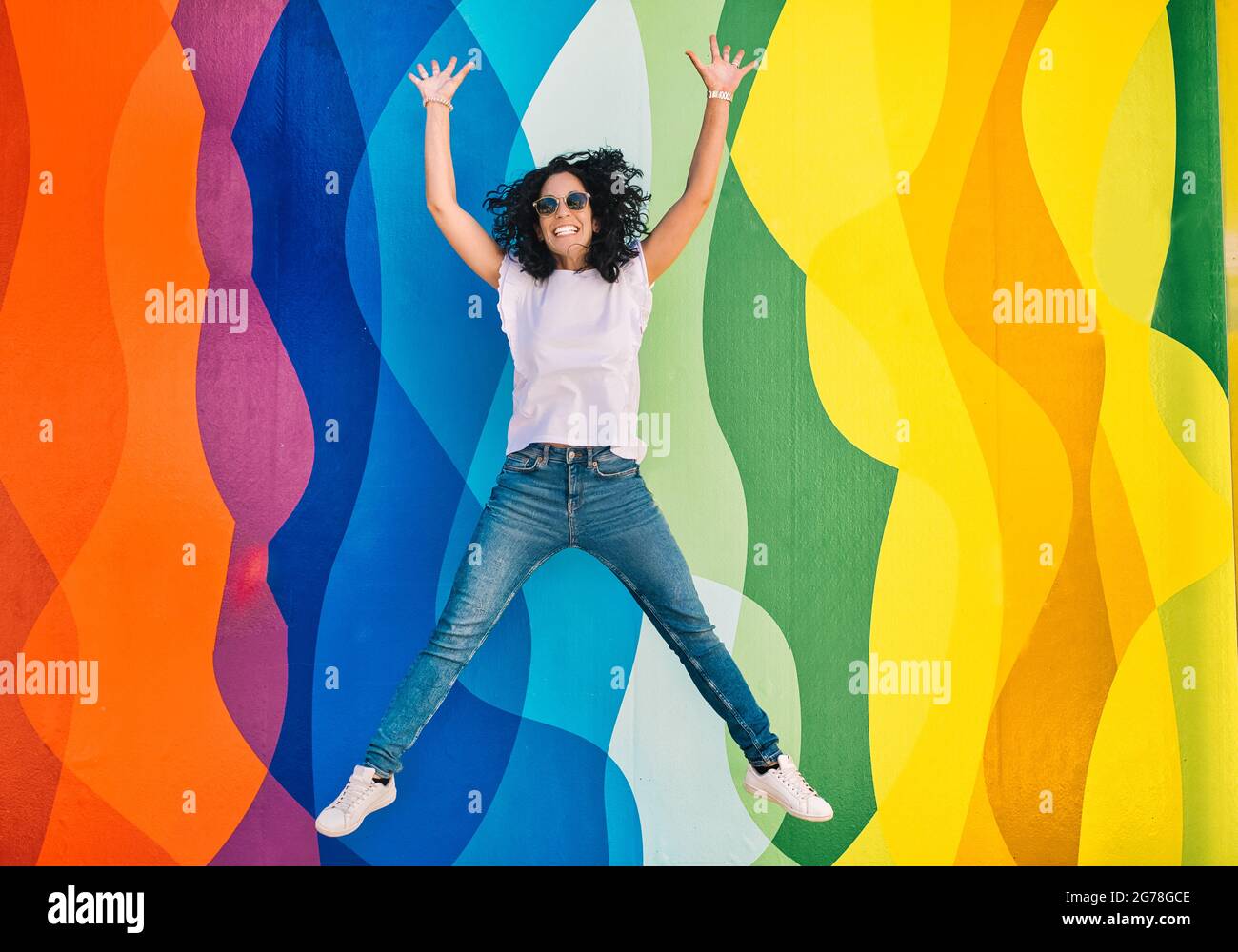 Young brunette woman with curly hair, jeans and sunglasses has fun while jumping on a colorful wall. leisure and free time concept Stock Photo