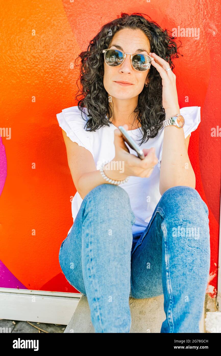 Portrait of a young pretty woman with curly black hair and sunglasses smiling while sitting on the street with her mobile phone with a red wall in the Stock Photo