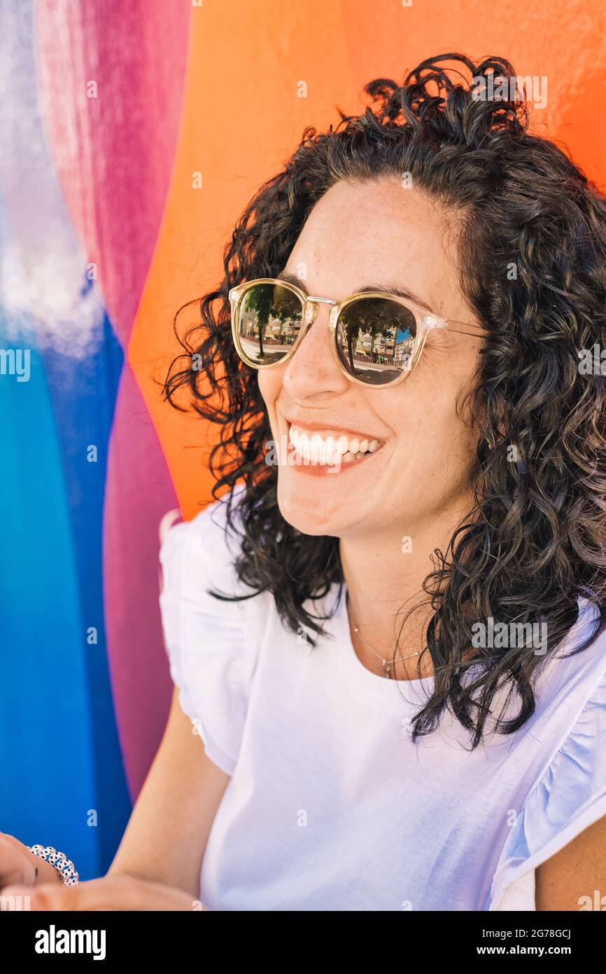 portrait of a young woman with curly black hair and sunglasses laughing in the street on a background colorful wall Stock Photo