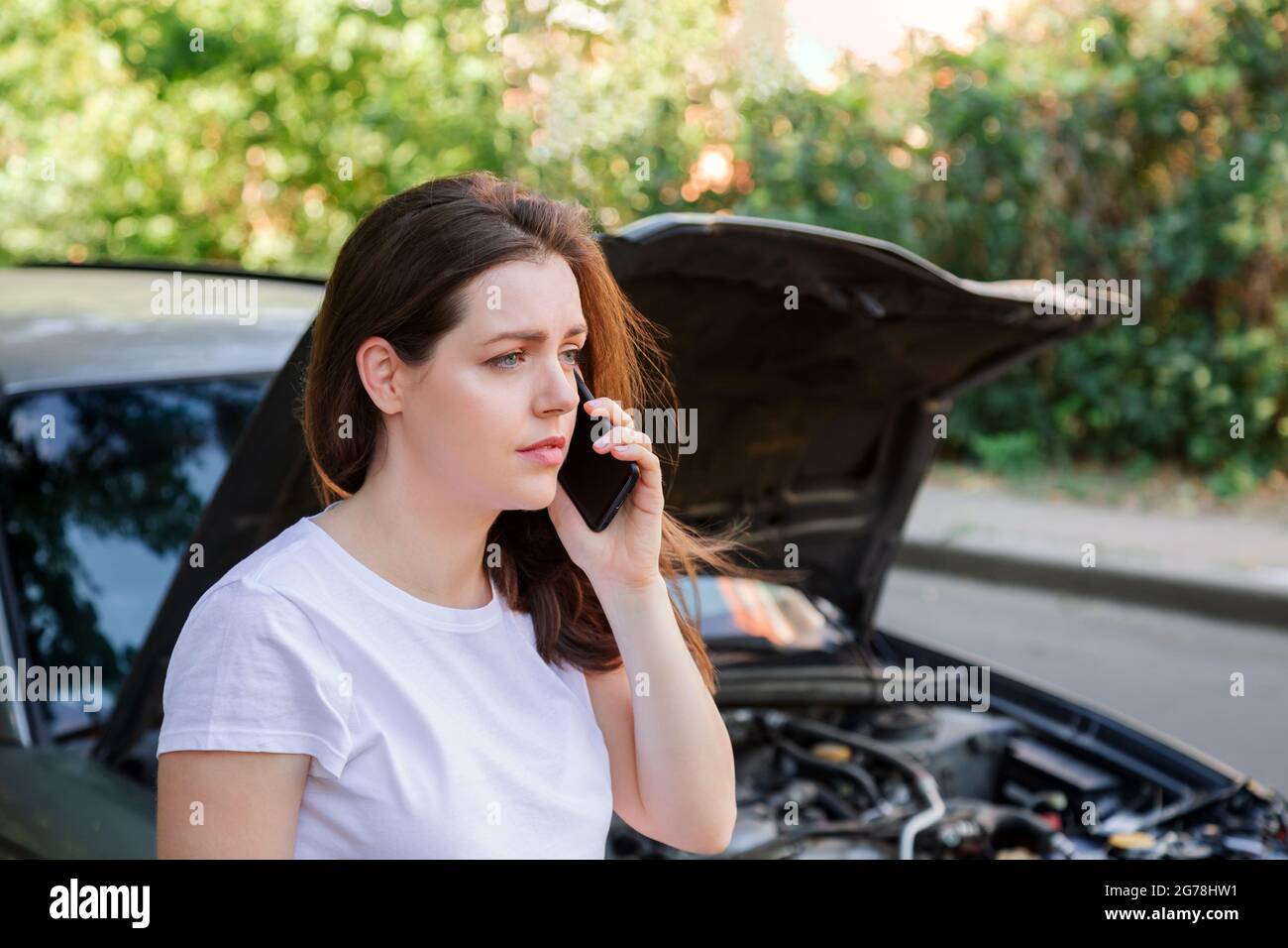Portrait of frustrated young woman calling mobile phone for help Insurance service or ambulance after car accident. Woman in front of broken car with Stock Photo