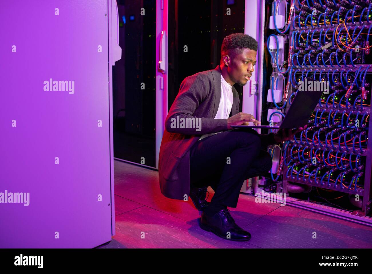 Busy young black datacenter engineer crouching in server room with neon illumination and checking network settings on laptop Stock Photo
