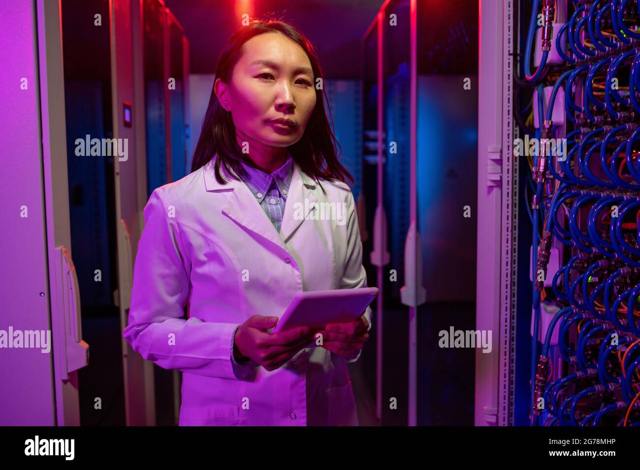 Portrait of serious young Asian support technician in white coat standing with tablet in dark neon-illuminated room Stock Photo