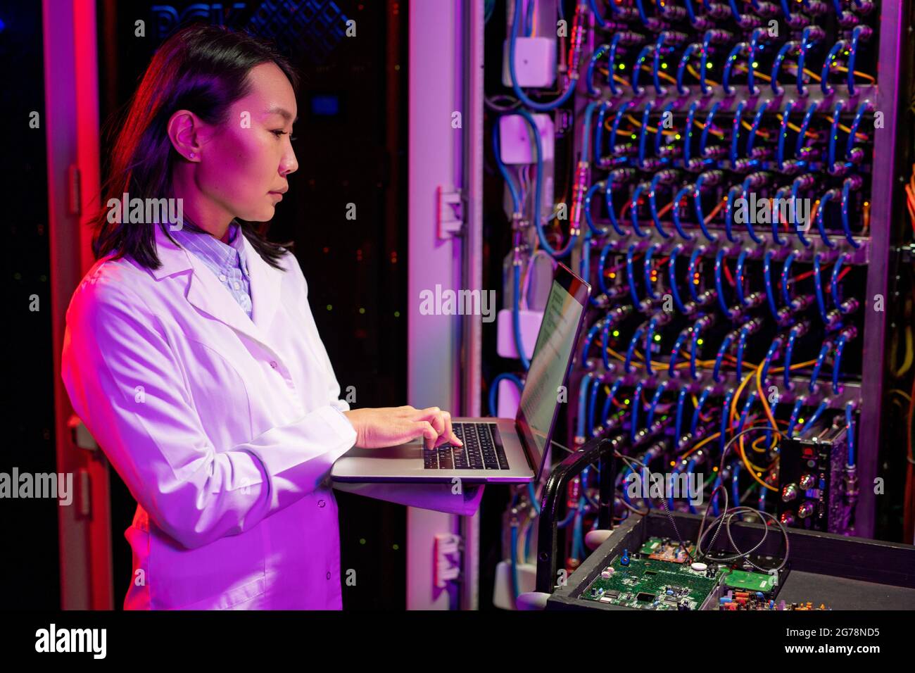 Serious busy young Asian female network engineer in lab coat standing at cart with circuit board connected to server and analyzing data on laptop Stock Photo