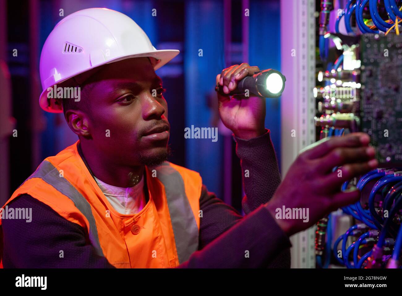 Serious young African-American server repairman in hardhat using flashlight while checking server rack Stock Photo