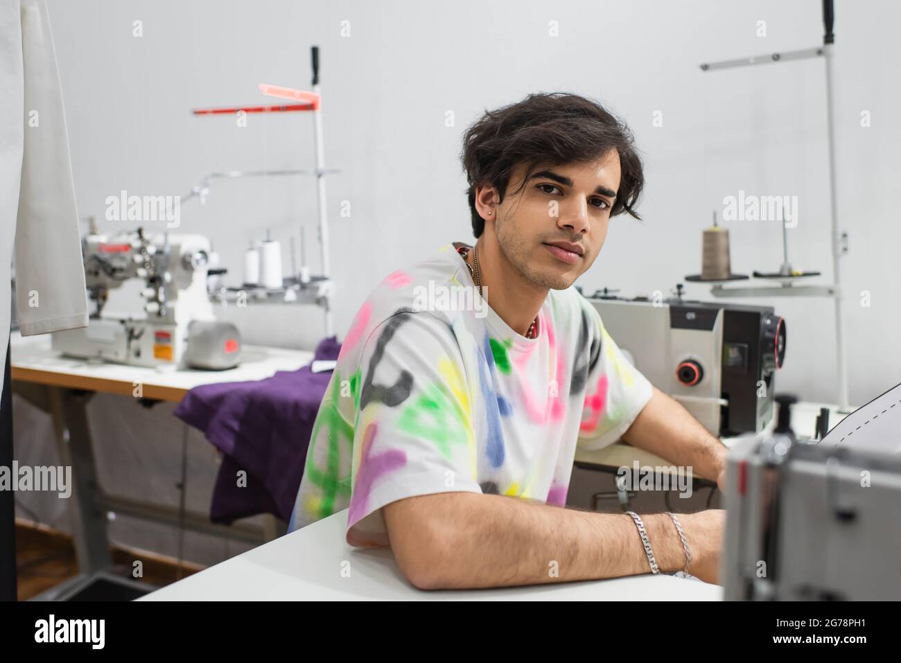 stylish tailor looking at camera while working on sewing machine in ...