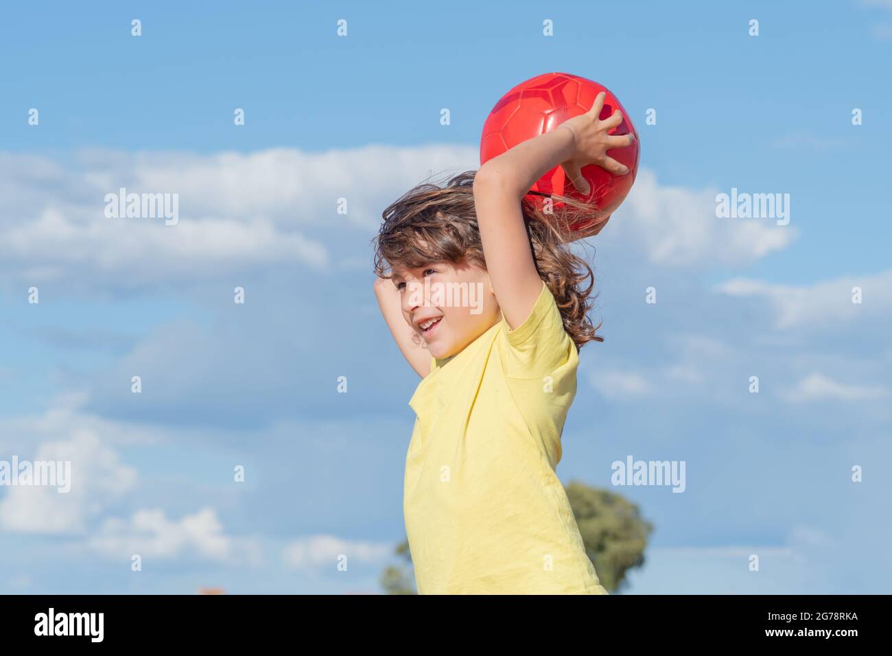 little boy with long hair and yellow t-shirt plays with his red ball in a field on a sunny summer day Stock Photo