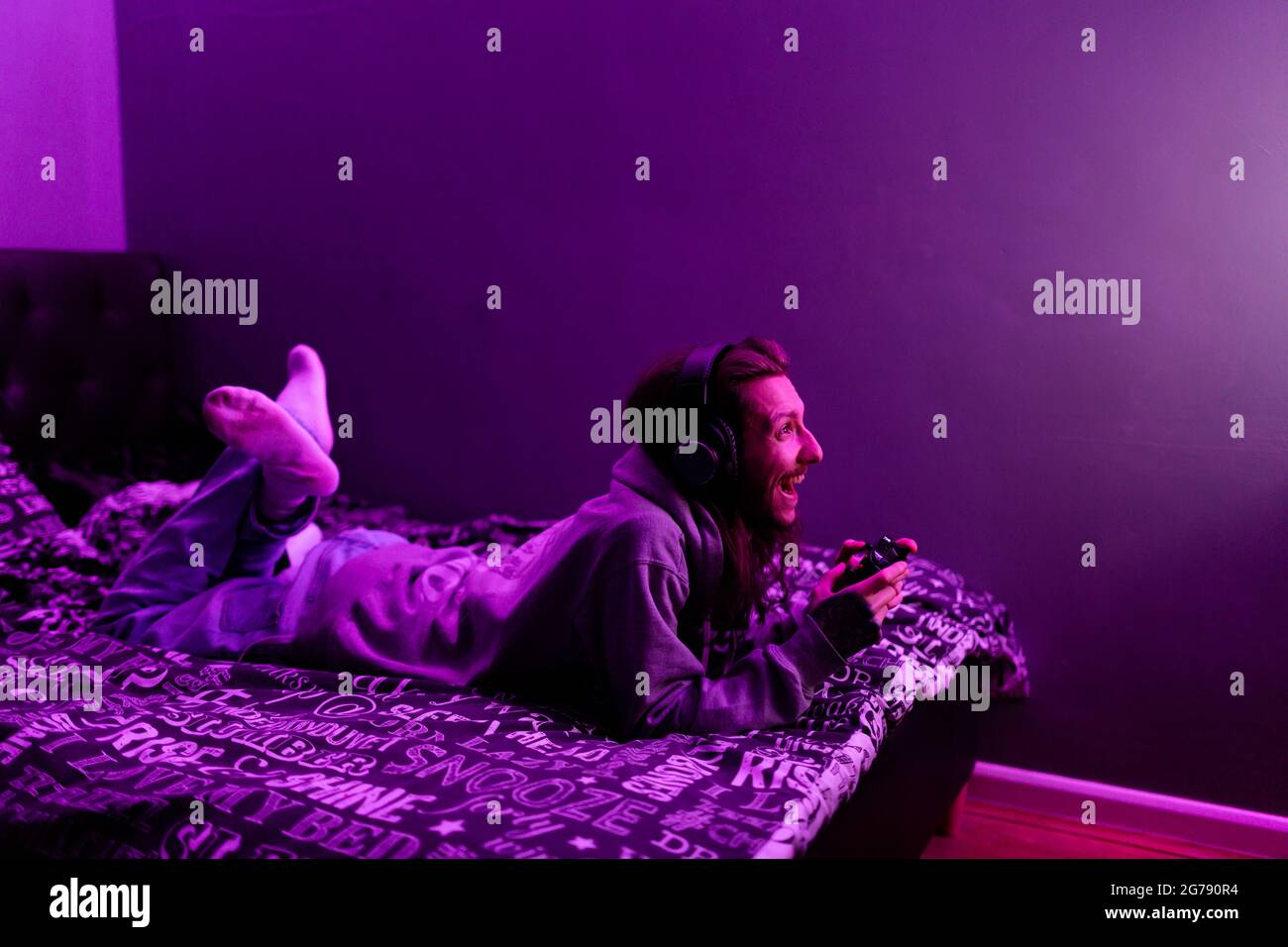 A young man sitting on his bed playing video games, he has the lights on and his lit up by the TV. After dark concept Stock Photo
