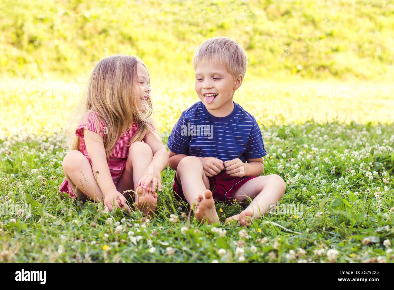 A blonde-haired boy and a girl are sitting on a green grass lawn and playing with each other. Happy childhood, fun vacation Stock Photo