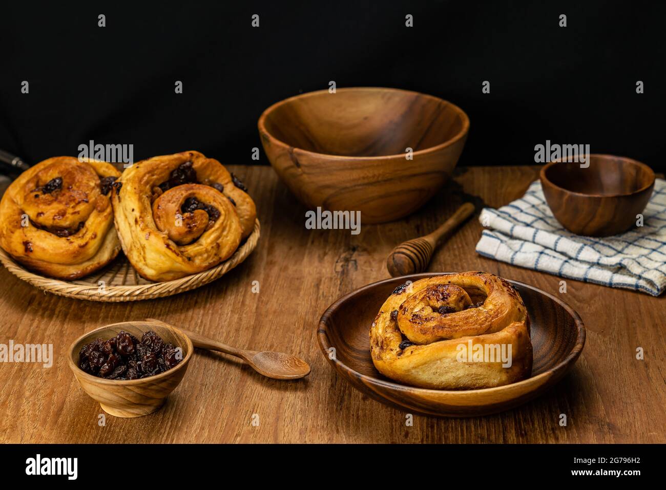 Homemade delicious raisin cinnamon cake roll in wooden plate and in bamboo tray with dried raisin in wooden cup, spoon, honey dipper, bowl and table c Stock Photo