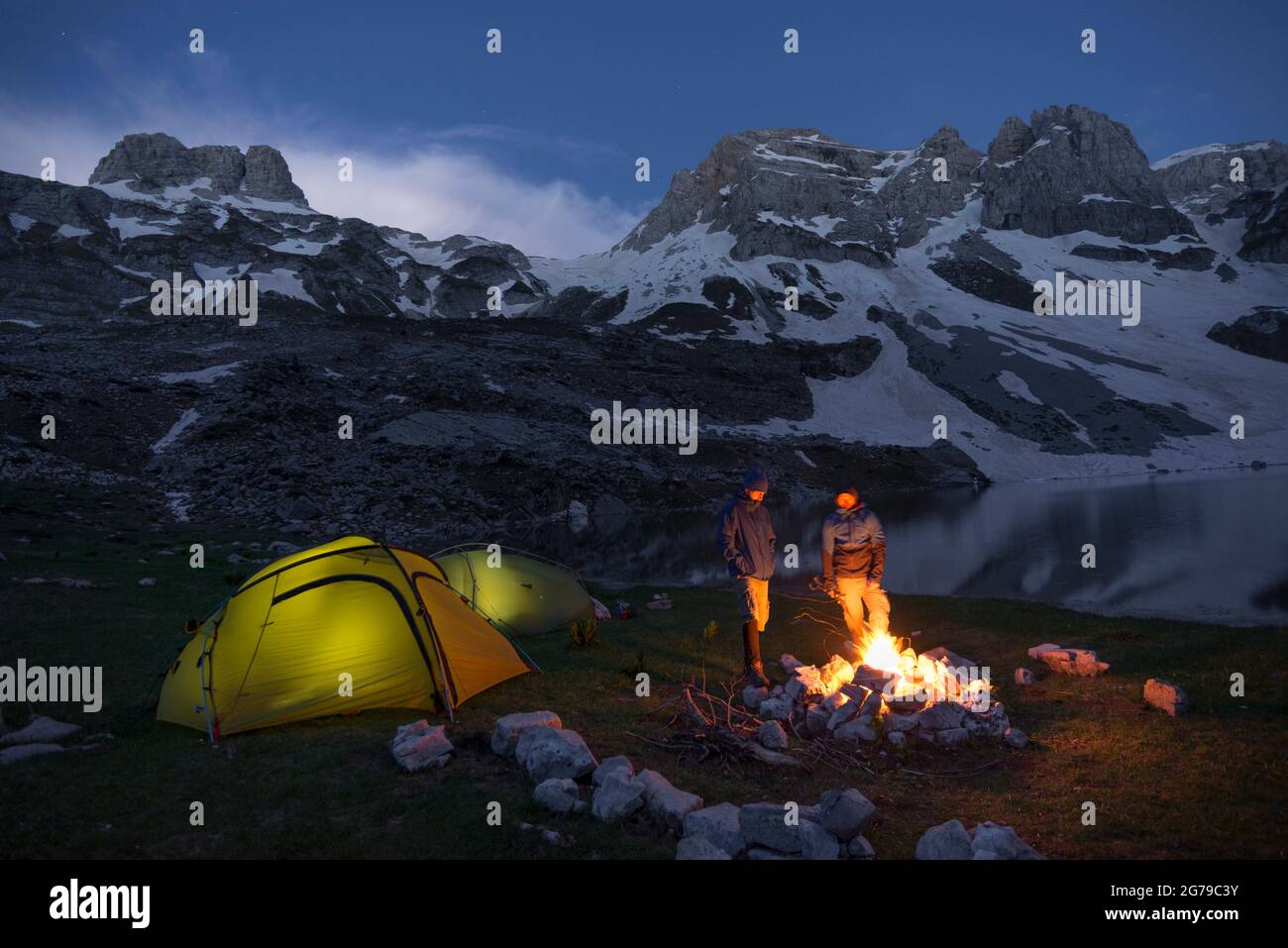 Two hikers stand by a campfire next to their lighted tents in the Prokletije National Park near a mountain lake, Albania Stock Photo