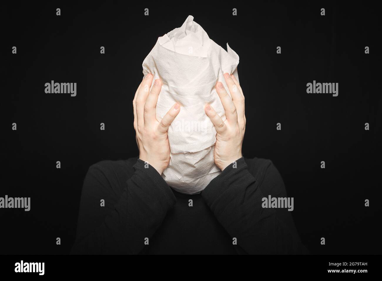 Head wrapped in toilet paper. Man holds his hands behind his head. Funny man, black background. Mummy, mental health concept. Oh my god. Stock Photo