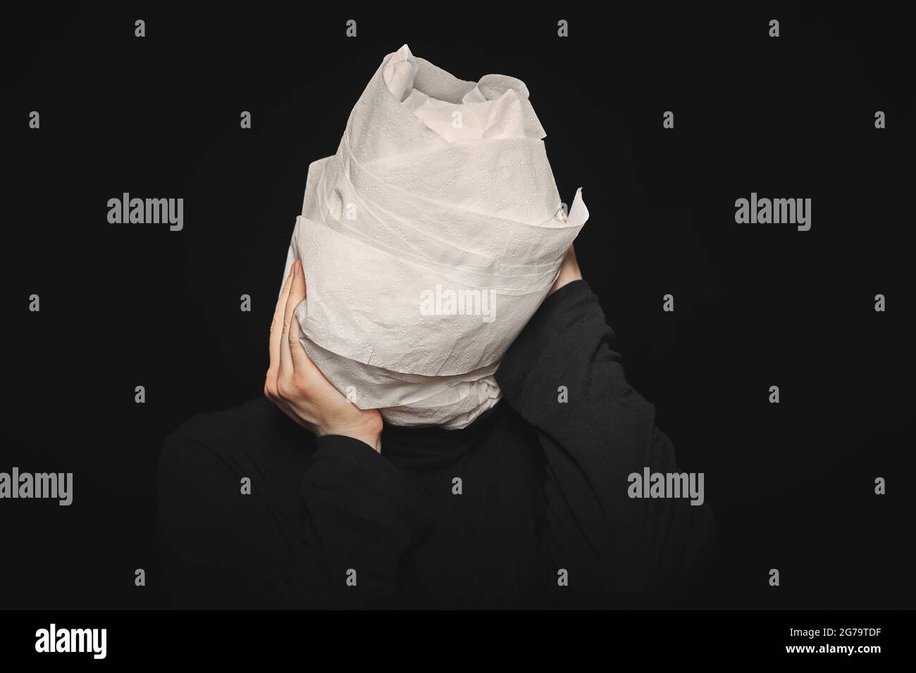 Head wrapped in toilet paper. Man holds his hands behind his head. Funny man, black background. Mummy, mental health concept. Oh my god. Stock Photo