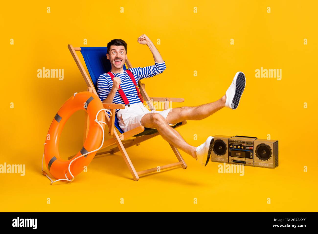Full body photo of happy victorious young man sit chair beach raise fists isolated on yellow color background Stock Photo