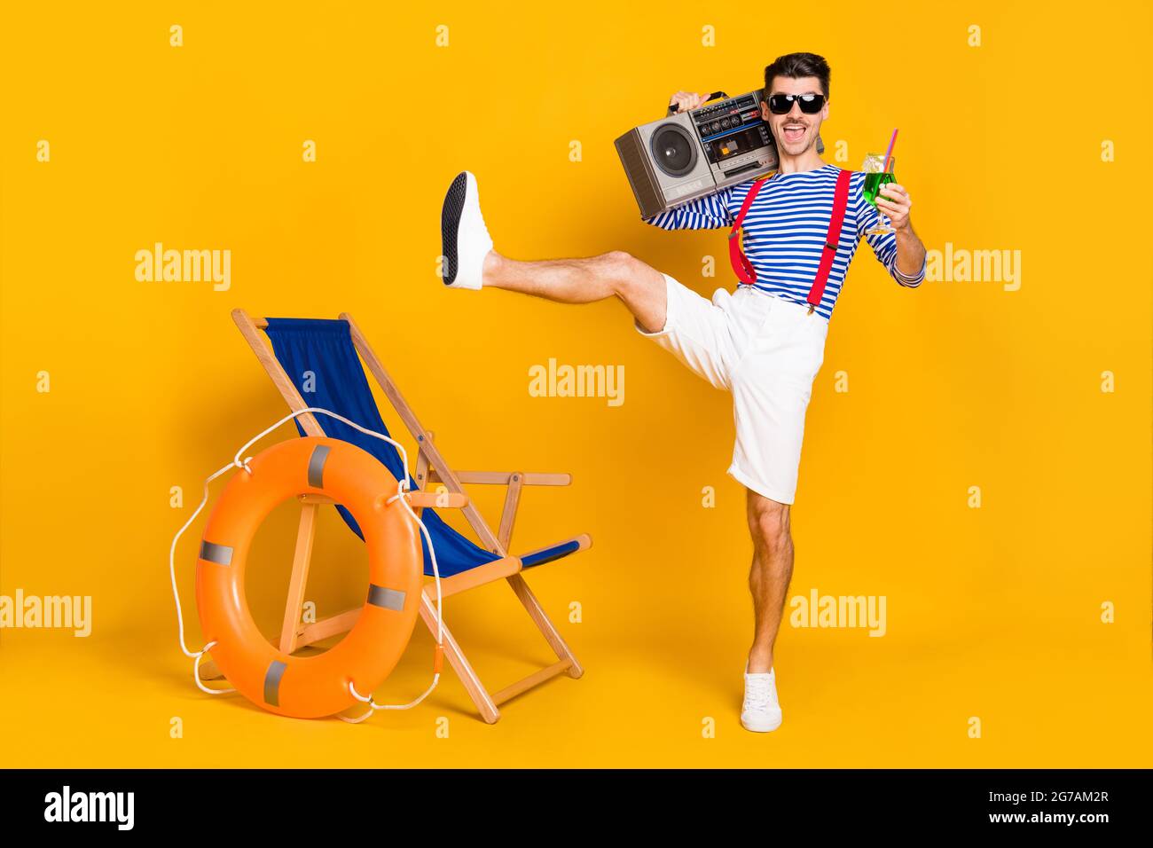 Full size photo of funky young brunette man wear red suspenders white shorts hold boombox drink beach isolated on yellow color background Stock Photo