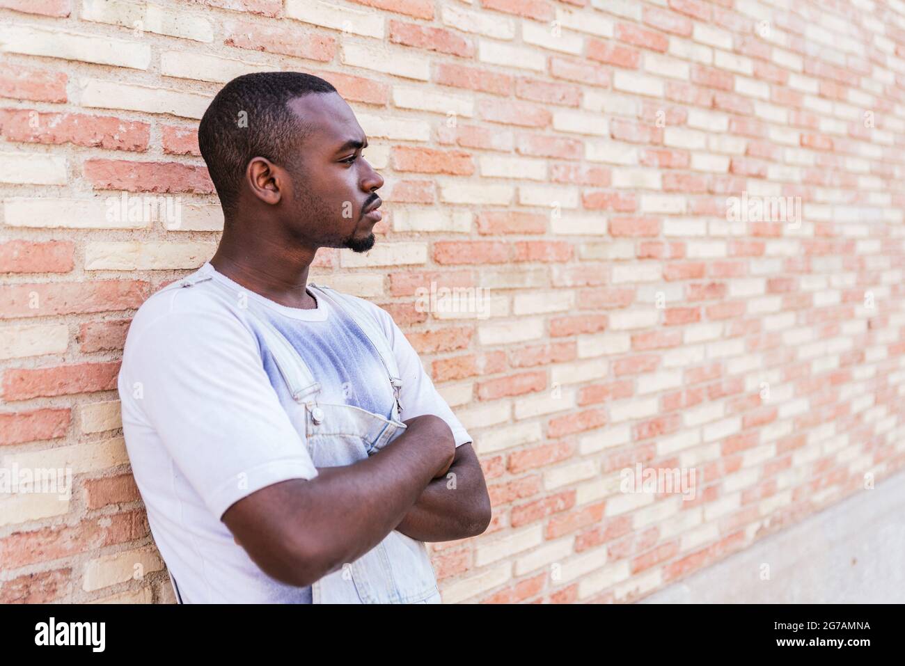 young afro man leaning on wall looking to one side on the streets. He looks serious or angry and crosses his arms Stock Photo