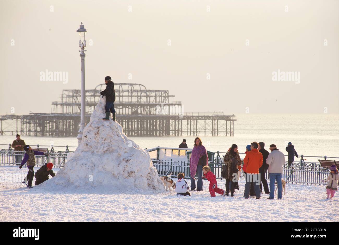Winter by the seaside. Kids and adults in the snow, building a snow 'stupa' snowman on Hove Lawns ... Brighton's dilapidated West Pier beyond. Brighton, England. Stock Photo