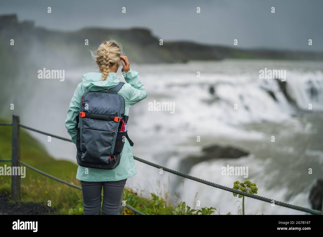 Blomd woman with backpack and green jacket visit Gullfoss powerful famous waterfall in Iceland. Famous place to visit on Golden Circle route of Iceland Stock Photo