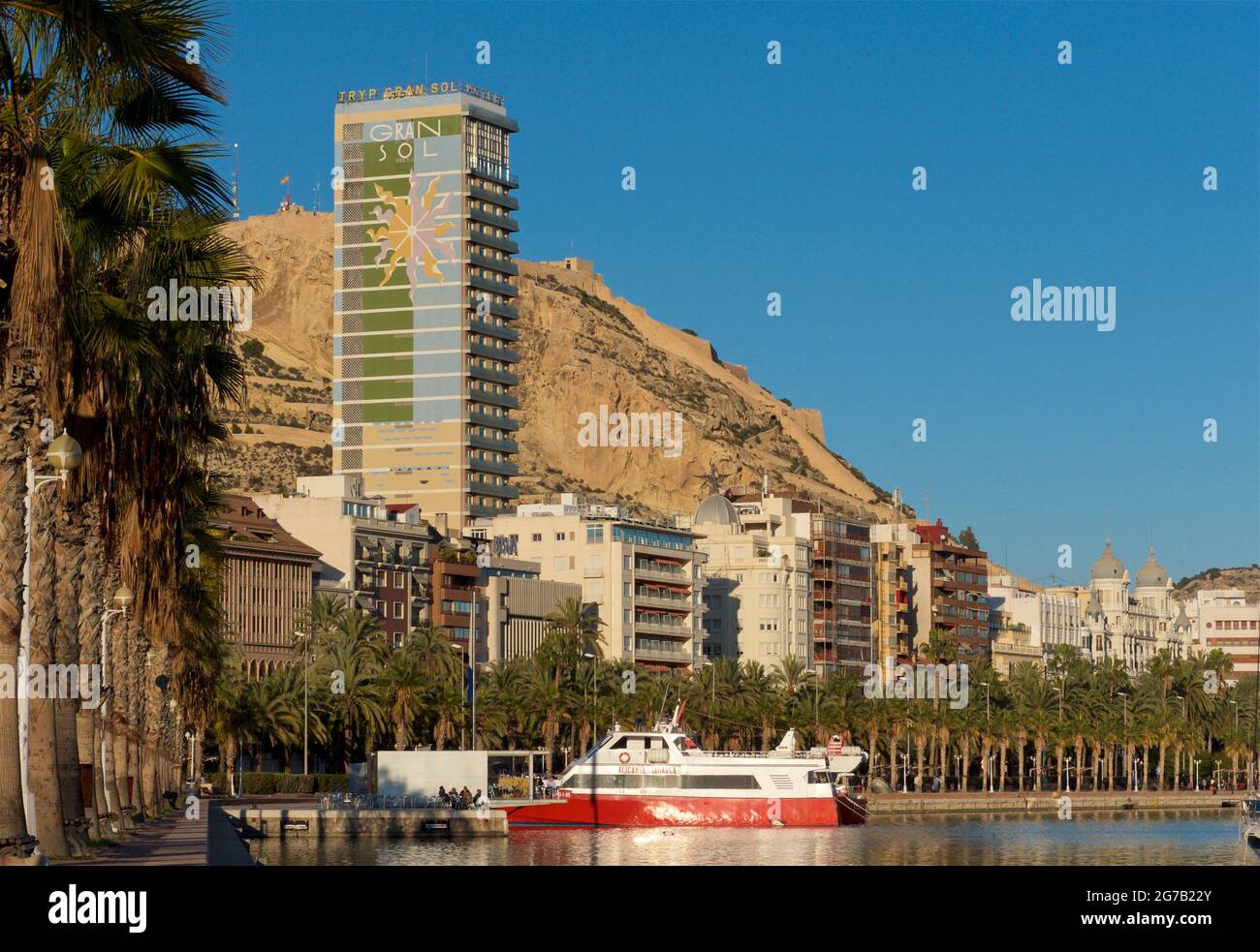 Palm-tree-lined Alicante promenade alongside the marina, Alicante, Valencia, Spain. The 97m high Gran Sol building beyond. Also known as Hotel Gran Sol, Hotel Tryp Gran Sol and officially Edificio Alonso. Mount Benacantil in the background Stock Photo
